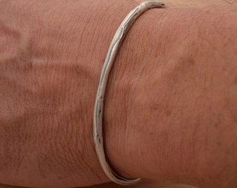 May include: A silver bangle bracelet with a hammered finish. The bracelet is worn on a person's wrist.