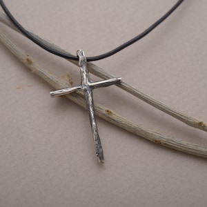 Può includere: Un ciondolo a forma di croce in argento con una superficie ruvida e testurizzata appeso a un cordino in pelle nera. Il ciondolo ha la forma di una croce semplice con un aspetto leggermente irregolare e rustico.