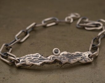 Könnte beinhalten: Ein silbernes Kettenarmband mit einem strukturierten, organisch geformten Silberstab, der in der Mitte einen schwarzen Diamanten trägt.