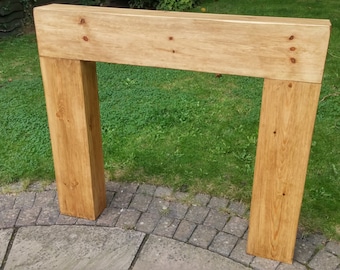 May include: A rectangular wooden fireplace mantel with a natural finish. The mantel is constructed from thick, solid wood beams, featuring visible wood grain and knots. It stands upright on a stone patio with a green lawn in the background.