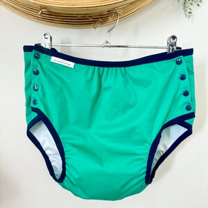 May include: Green cloth diaper with navy blue trim and snap closures. The diaper has a white interior and is hanging on a metal hanger. The brand name "branches of me" is printed on a small white tag.