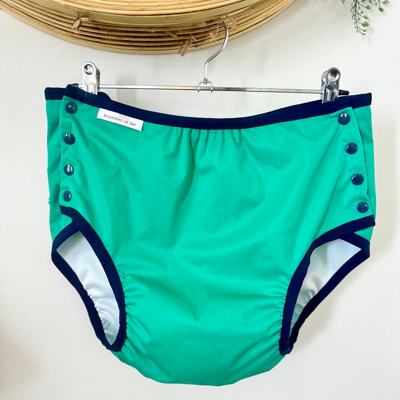 May include: Green cloth diaper with navy blue trim and snap closures. The diaper has a white interior and is hanging on a metal hanger. The brand name "branches of me" is printed on a small white tag.