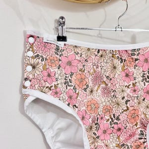 May include: Floral print underwear with a white trim and button details. The underwear features a pattern of pink, peach, and cream flowers on a light pink background. The garment is hanging on a metal hanger.