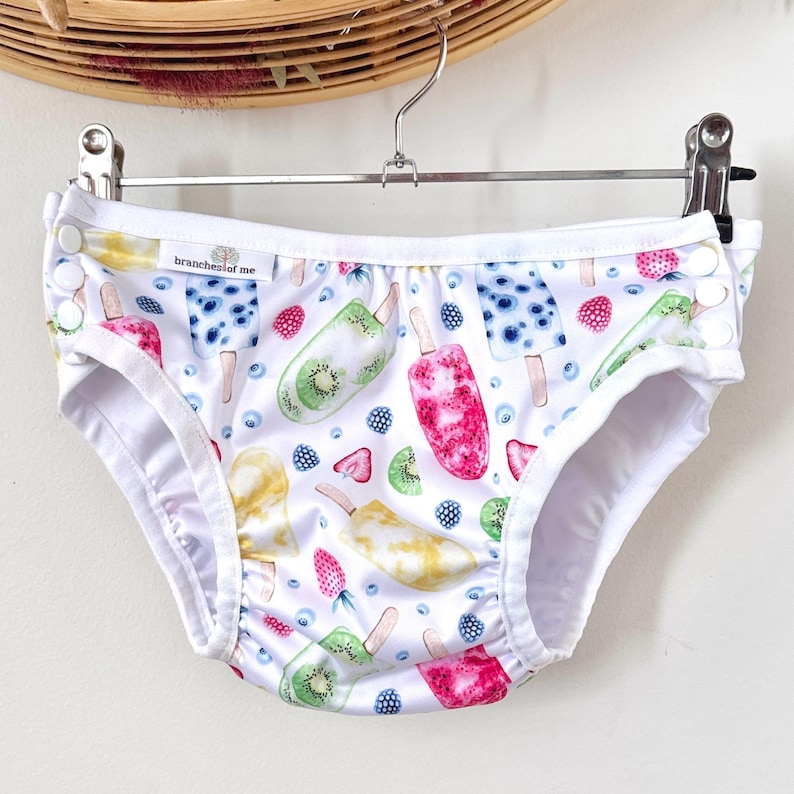 May include: White baby underwear with a colorful popsicle and fruit pattern. The design includes illustrations of kiwi, strawberry, and blueberry popsicles. The text "branches of me" is visible on the garment. The underwear is hanging on a metal hanger.