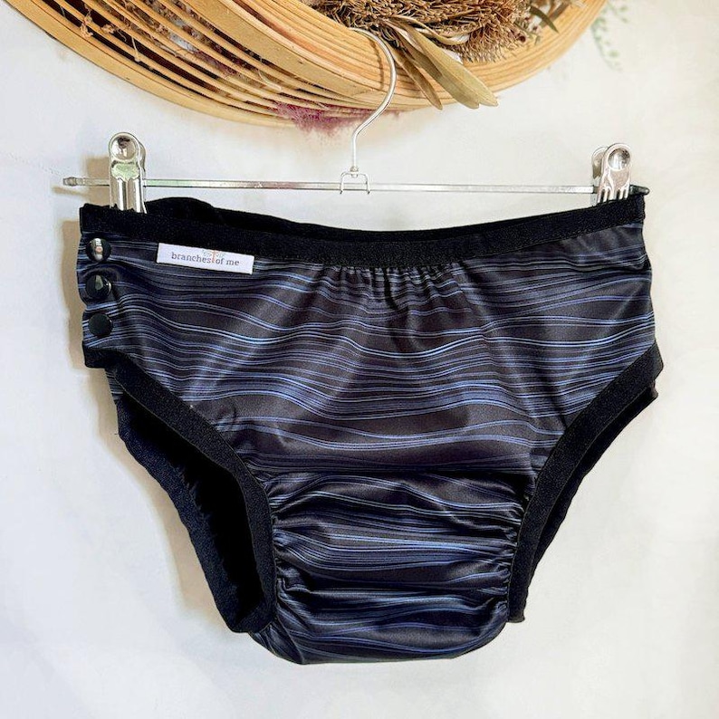 May include: Black underwear with a wavy blue and black striped pattern. The underwear has a black trim and a ruched front. The text "branchesofme" is printed on a small white tag. The underwear is hanging on a metal hanger.