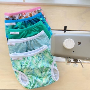 May include: A stack of colorful baby underwear with various patterns, including floral, abstract, and dinosaur prints. Each pair has a white trim and the text "branches of me". The underwear is displayed on a light wooden surface.