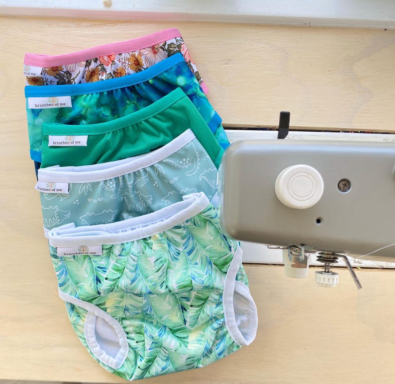 May include: A stack of colorful baby underwear with various patterns, including floral, abstract, and dinosaur prints. Each pair has a white trim and the text "branches of me". The underwear is displayed on a light wooden surface.