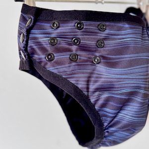 May include: A reusable cloth diaper with a dark blue and black striped pattern. The diaper has black snaps and a black elastic waistband. The design includes a series of snaps for adjustable sizing.