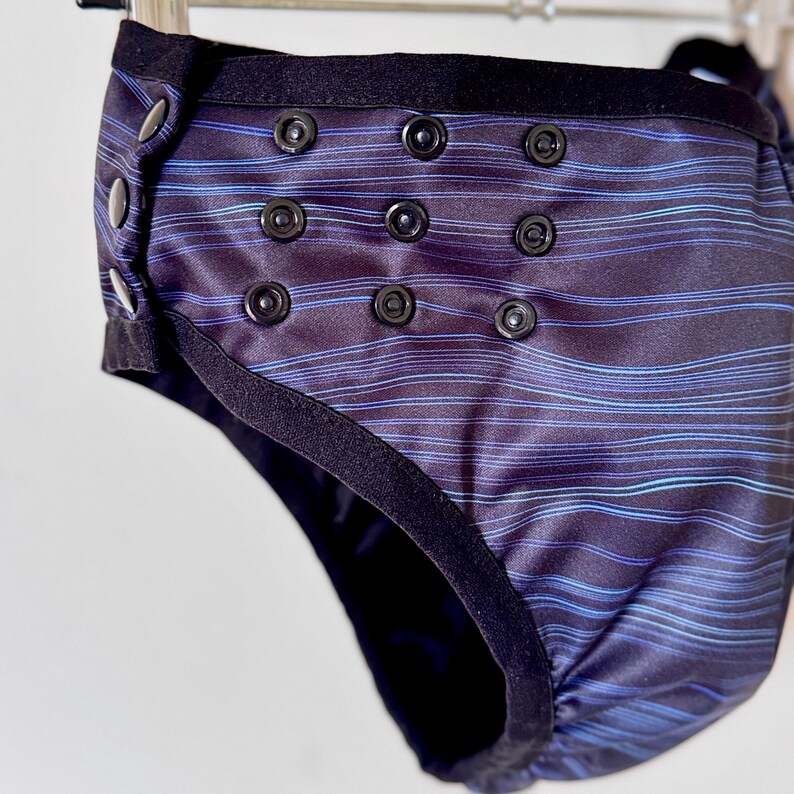 May include: A reusable cloth diaper with a dark blue and black striped pattern. The diaper has black snaps and a black elastic waistband. The design includes a series of snaps for adjustable sizing.