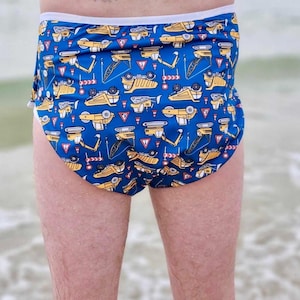 May include: Blue swim briefs with a repeating pattern of yellow construction vehicles and road signs. The briefs have a white waistband and trim. The image shows the briefs from the back, with the person's legs visible.