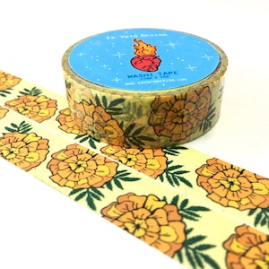 May include: A roll of yellow washi tape with a floral pattern of orange marigolds and green leaves. The tape is on a white background. The tape dispenser is blue with a white label that says "EX VOTO DESIGN" and "WASHITAPE 15mm x 10m" and a graphic of a flaming heart.