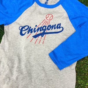 Pode incluir: Uma camiseta de beisebol cinza e azul com a palavra "Chingona" em uma fonte azul de escrita com uma bola de beisebol e raios vermelhos atr&aacute;s.