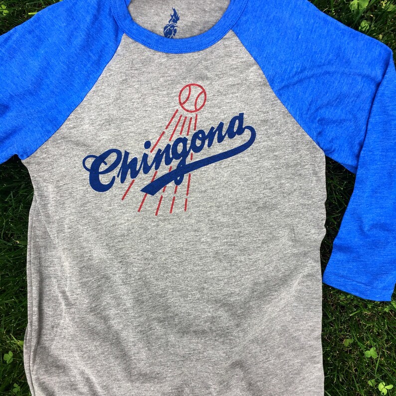 Pode incluir: Uma camiseta de beisebol cinza e azul com o texto "Chingona" em uma fonte azul cursiva com uma bola de beisebol vermelha e raios vermelhos atr&aacute;s.