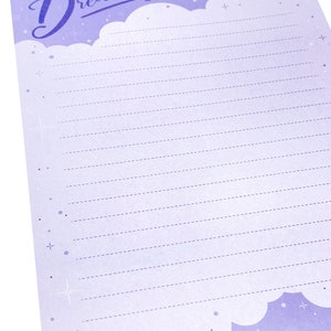May include: A lavender-colored notepad with the words "dreaming of..." in a script font. The notepad has a cloud design at the top and bottom, with dotted lines for writing. Small stars and dots are scattered across the page.