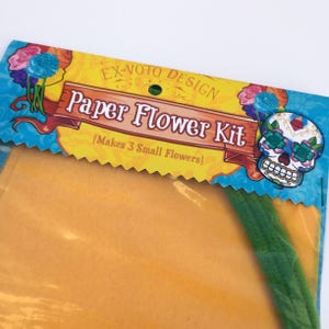 K&ouml;nnte beinhalten: Eine gelbe und blaue Verpackung mit dem Text "Ex-Voto Design Paper Flower Kit (Makes 3 Small Flowers)" und einer bunten Zucker Sch&auml;del Illustration. Die Verpackung enth&auml;lt gelbes Seidenpapier und gr&uuml;ne Stiele zum Basteln von Papierblumen.