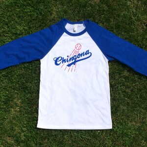 K&ouml;nnte beinhalten: Ein wei&szlig;es und blaues Baseball-T-Shirt mit einem roten Baseball und dem Text "Chingona" in Blau.