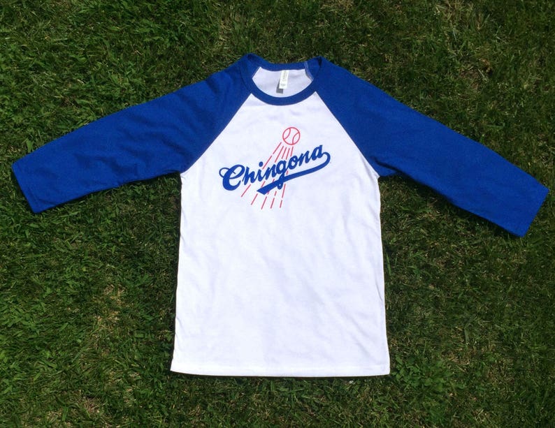 K&ouml;nnte beinhalten: Ein wei&szlig;es und blaues Baseball-T-Shirt mit einem roten Baseball und dem Text "Chingona" in Blau.