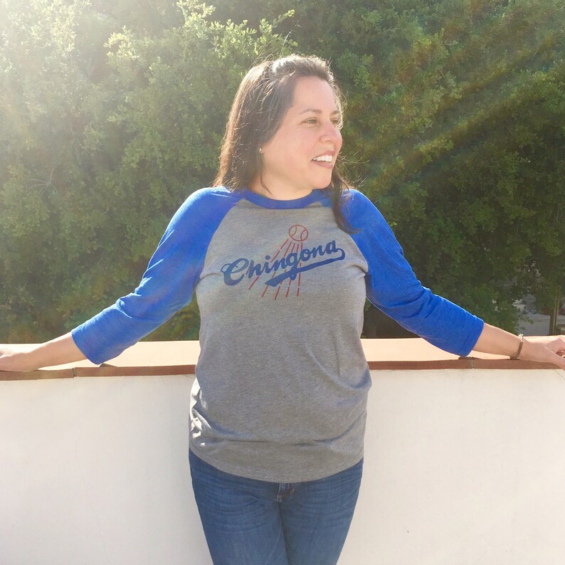 Pode incluir: Uma camiseta de estilo beisebol cinza e azul com o texto "Chingona" em letras azuis. A camiseta tem um esquema de cores azul e cinza e apresenta um gr&aacute;fico de taco de beisebol e bola.