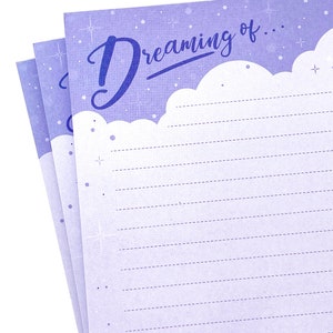 May include: A stack of lined notepaper with a light purple background and the words "Dreaming of..." in a script font. The top edge of the paper has a cloud-like design. The paper is decorated with small stars and dots.