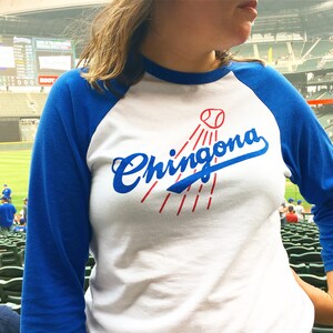 K&ouml;nnte beinhalten: Ein wei&szlig;es und blaues Baseball-T-Shirt mit einer blauen und roten Grafik, auf der "Chingona" steht, mit einer Baseball-Grafik &uuml;ber dem Wort.