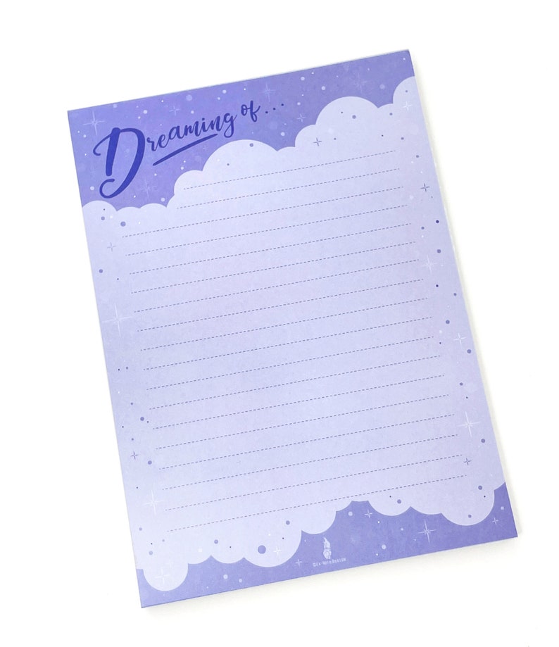 May include: A lavender-colored notepad with the words "Dreaming of..." in a script font. The top edge features a cloud design, and the lined paper is decorated with stars. The notepad is designed for writing and note-taking.