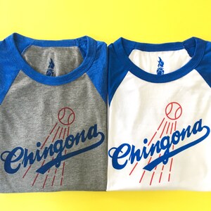 K&ouml;nnte beinhalten: Zwei Baseball-T-Shirts in einem blau-grauen Farbschema. Die Shirts zeigen den Text "Chingona" in einer stilisierten Schriftart mit einem Baseball und roten Strahlen, die von ihm ausgehen.