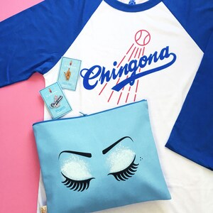 K&ouml;nnte beinhalten: Ein blaues und wei&szlig;es Baseballtrikot mit dem Wort "Chingona" auf der Vorderseite. Das Trikot wird mit einer hellblauen Rei&szlig;verschlusstasche mit einem wei&szlig;en Glitzeraugen-Design kombiniert.