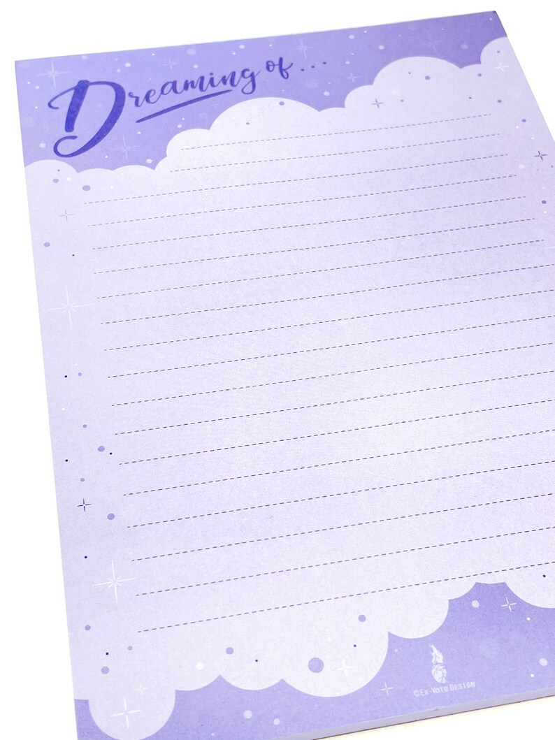 May include: A lavender-colored notepad with the words "dreaming of..." in a script font. The notepad has a cloud design at the top and bottom, with dotted lines for writing. Small stars and dots are scattered across the page.