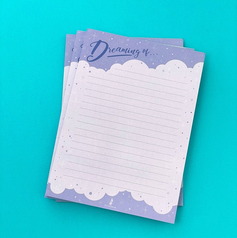 May include: A stack of lined notepads with a light purple cloud design border and the words "Dreaming of..." in a script font. The notepads have a light purple background with white lines and small star accents.