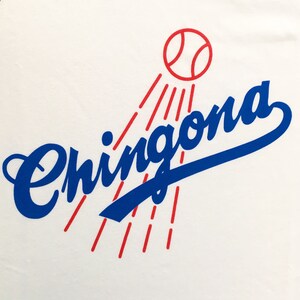 K&ouml;nnte beinhalten: Ein wei&szlig;es T-Shirt mit einem blauen und roten Grafikdesign. Die Grafik zeigt das Wort "Chingona" in blauer Kursivschrift mit einem roten Baseball dar&uuml;ber und roten Lichtstrahlen, die vom Baseball ausgehen.
