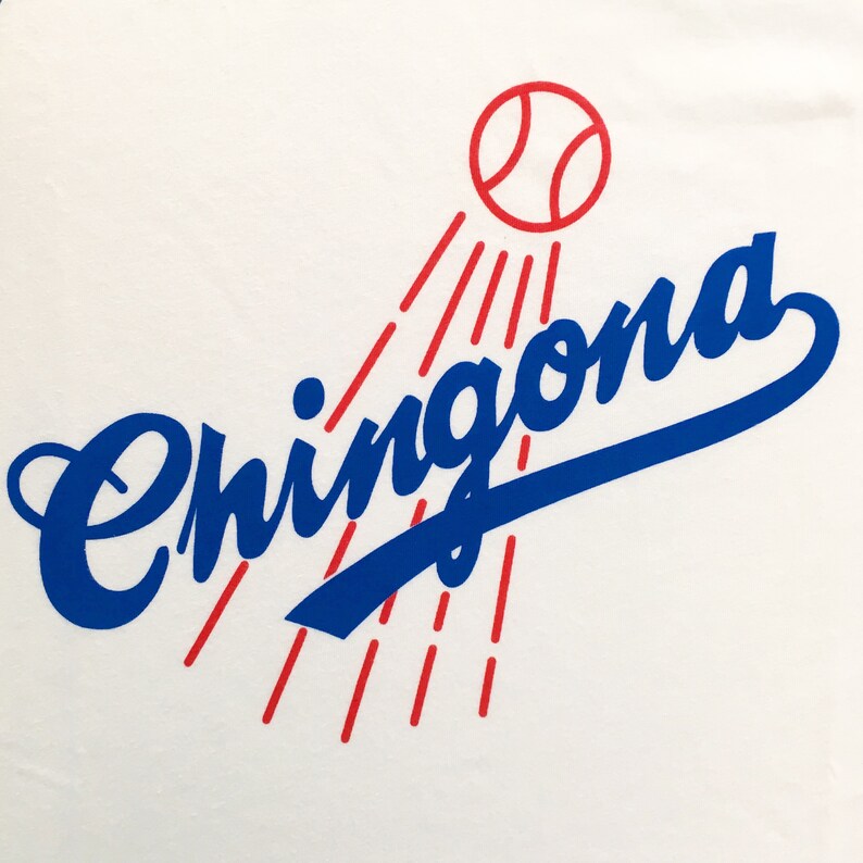 K&ouml;nnte beinhalten: Ein wei&szlig;es T-Shirt mit einem blauen und roten Grafikdesign. Die Grafik zeigt das Wort "Chingona" in blauer Kursivschrift mit einem roten Baseball dar&uuml;ber und roten Lichtstrahlen, die vom Baseball ausgehen.