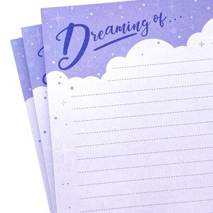 May include: A stack of lined notepaper with a light purple background and the words "Dreaming of..." in a script font. The top edge of the paper has a cloud-like design. The paper is decorated with small stars and dots.