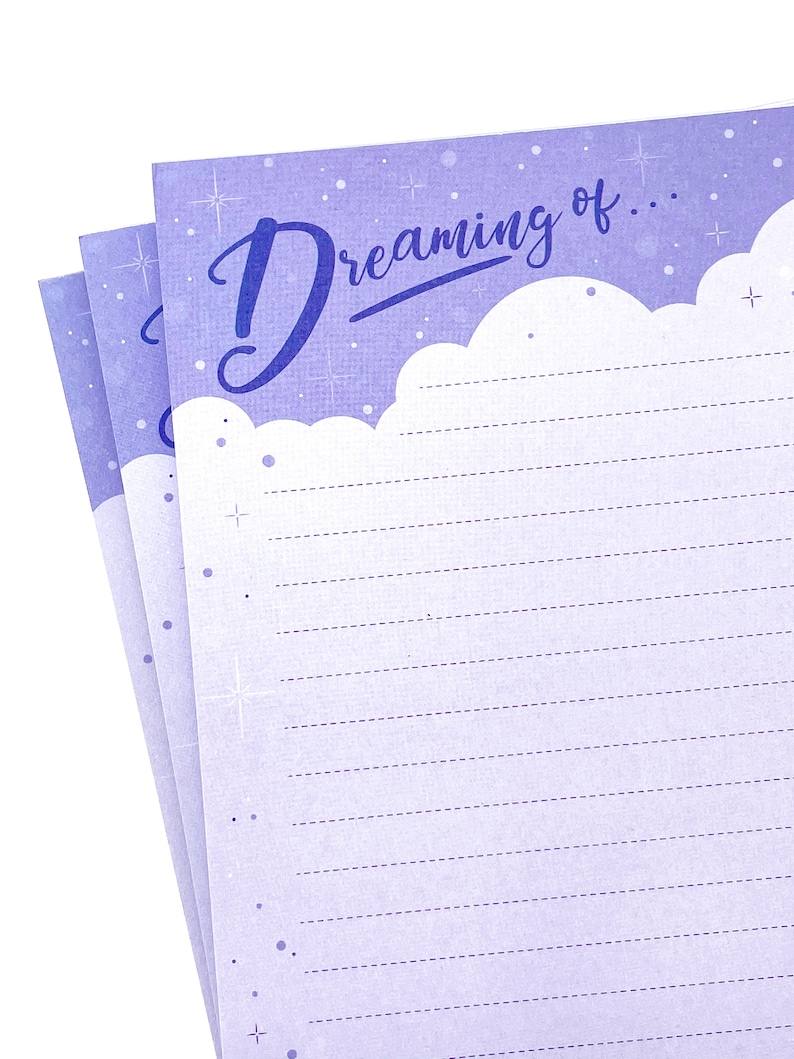 May include: A stack of lined notepaper with a light purple background and the words "Dreaming of..." in a script font. The top edge of the paper has a cloud-like design. The paper is decorated with small stars and dots.