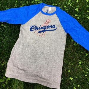 Pode incluir: Uma camiseta de beisebol cinza e azul royal com uma bola de beisebol vermelha e o texto "Chingona" em azul.
