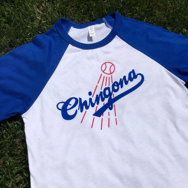K&ouml;nnte beinhalten: Ein wei&szlig;es und blaues Baseball-T-Shirt mit einem roten Baseball und dem Wort "Chingona" in blauer Kursivschrift.