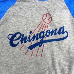 Pode incluir: Uma camiseta de beisebol cinza e azul com a palavra "Chingona" em letras cursivas azuis e uma bola de beisebol vermelha com costuras brancas acima da palavra.
