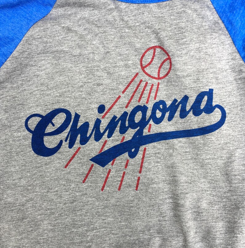 Pode incluir: Uma camiseta de beisebol cinza e azul com a palavra "Chingona" em letras cursivas azuis e uma bola de beisebol vermelha com costuras brancas acima da palavra.
