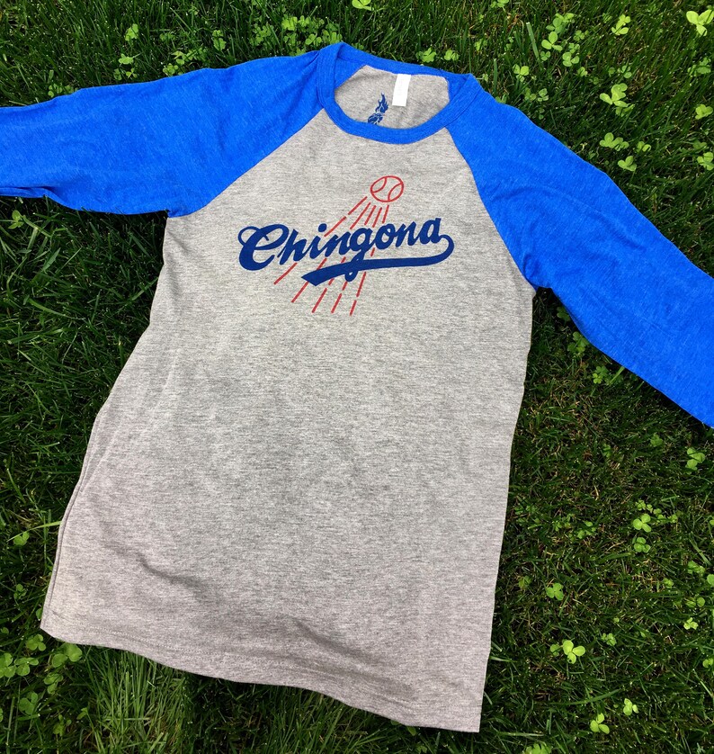Pode incluir: Uma camiseta de beisebol cinza e azul royal com uma bola de beisebol vermelha e o texto "Chingona" em azul.
