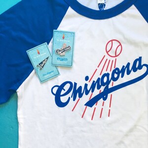 K&ouml;nnte beinhalten: Ein wei&szlig;es Baseball-T-Shirt mit einem blauen und roten Grafikdesign, auf dem "Chingona" mit einem Baseball und roten Lichtstrahlen steht. Auf dem Shirt befinden sich zwei Emaille-Pins, einer sagt "Chingona" und der andere "Los Angeles".