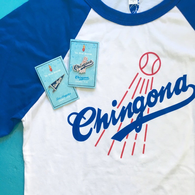 K&ouml;nnte beinhalten: Ein wei&szlig;es Baseball-T-Shirt mit einem blauen und roten Grafikdesign, auf dem "Chingona" mit einem Baseball und roten Lichtstrahlen steht. Auf dem Shirt befinden sich zwei Emaille-Pins, einer sagt "Chingona" und der andere "Los Angeles".