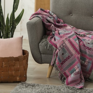 May include: A grey sofa with a pink and grey patterned throw blanket draped over the back. The sofa is in a living room setting with a potted plant and a wicker basket with a pink cushion.
