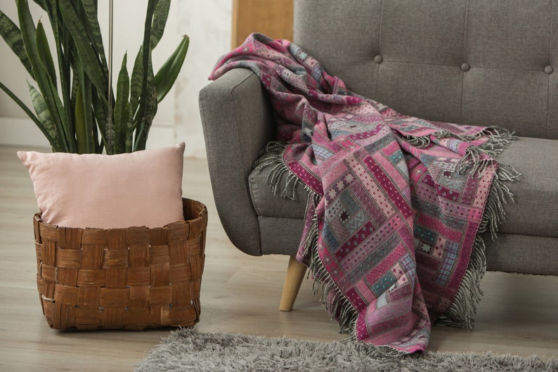 May include: A grey sofa with a pink and grey patterned throw blanket draped over the back. The sofa is in a living room setting with a potted plant and a wicker basket with a pink cushion.