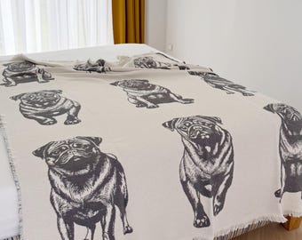 May include: A cream-colored woven throw blanket featuring a repeating pattern of dark gray pug dogs in various poses. The blanket has a textured fabric finish and a fringed edge, draped across a bed surface with a visible white and blue striped base.