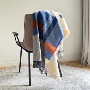 May include: A woven throw blanket featuring a plaid pattern in shades of blue, cream, burnt orange, and grey. The blanket has a soft texture and is finished with fringe detailing along the edges, draped over a modern chair with black metal legs.