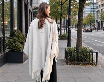 May include: A light gray, oversized poncho featuring a textured knit fabric, a draped silhouette, and long fringe detailing along the bottom hem. The garment is worn over black trousers, showcasing a neutral color palette and a soft, woven appearance.