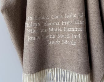 May include: A light brown blanket with a herringbone pattern and white fringe at the bottom. The fabric features white embroidered text listing names including Max, Louisa, Clara, Isalie, Philipp, Johanna, Fritz, Nikolaus, Marie, Hemma, Jonas, Janina, Matti, Jari, Jakob, and Nicole.