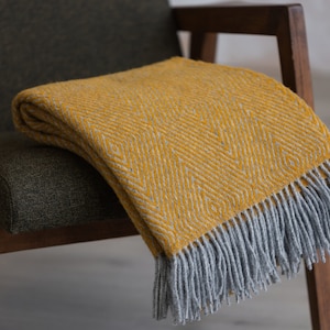 以下が含まれることがあります： 幾何学模様とグレーのフリンジが付いたマスタードイエローのブランケットが、濃い緑色の肘掛け椅子の上に置かれています。ブランケットの模様は、明るい黄色の繰り返しひし形で、質感のある外観を作り出しています。椅子は木製のフレームです。