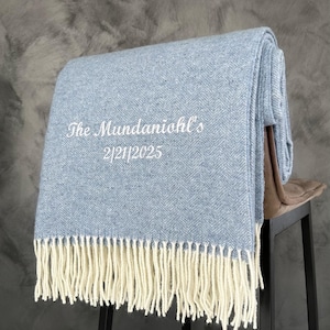 May include: A light blue throw blanket with a herringbone pattern and white fringe. The blanket is folded and draped over a brown stool. The text "The Mundaniol's 2/21/2025" is embroidered on the blanket.