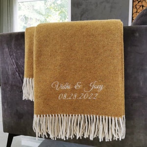Op de afbeelding: Mosterdkleurige plaid met witte franjes, gedrapeerd over een grijze bank. De plaid heeft de geborduurde tekst "Vidhi & Joey" en de datum "08.28.2022". De plaid is gemaakt van een textuurstof.