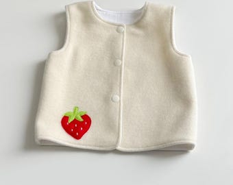 May include: A sleeveless cream-colored vest featuring a round neckline and a front button closure with three white snaps. The garment is decorated with a single red strawberry appliqué with a green stem positioned on the lower left side.